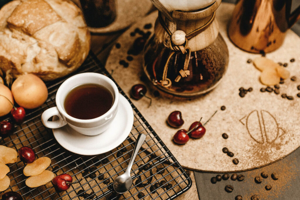 Cup of black coffee beside a pour-over carafe, bread, and cherries.